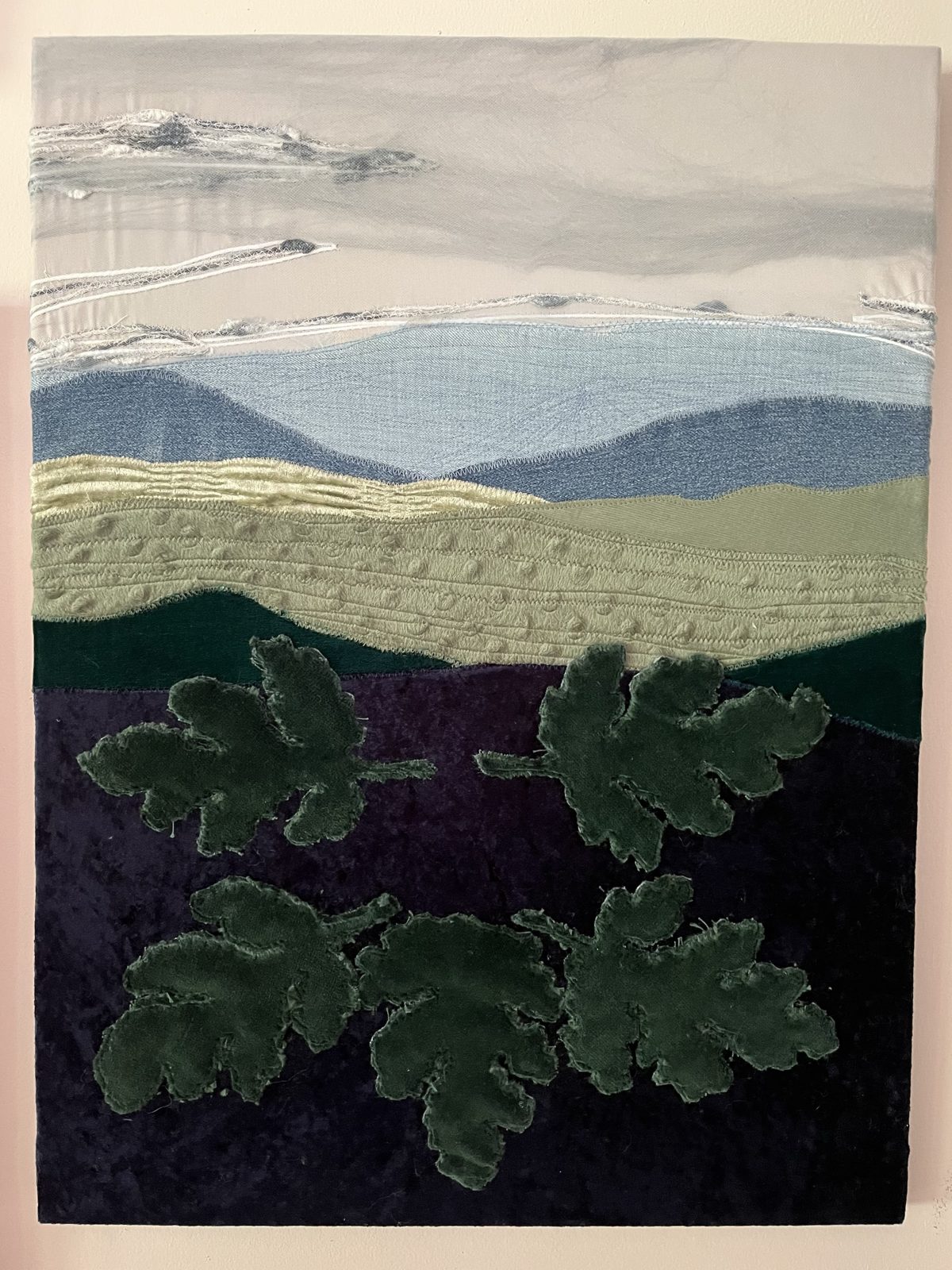 Vertical landscape quilt: couched white clouds, denim-blue ridges, green hills and dark green velvet oak leaves over a deep purple foreground