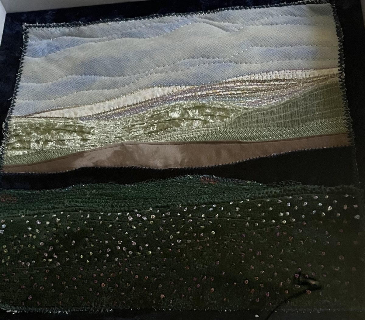 Small landscape quilt in a white shadow box on dark velvet, with a hand-dyed sky, couched ridges and French knots on a dark green hill