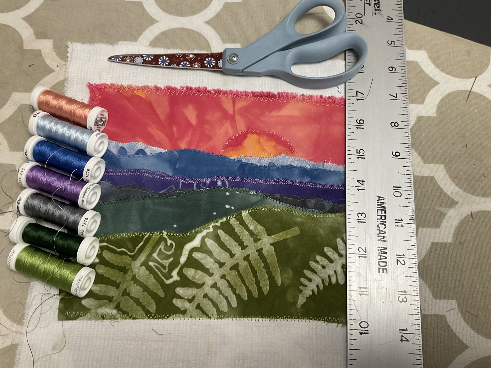 A sunset landscape in progress on the cutting table, raw-edge layers zigzagged down, with seven spools of rayon thread, scissors and a ruler