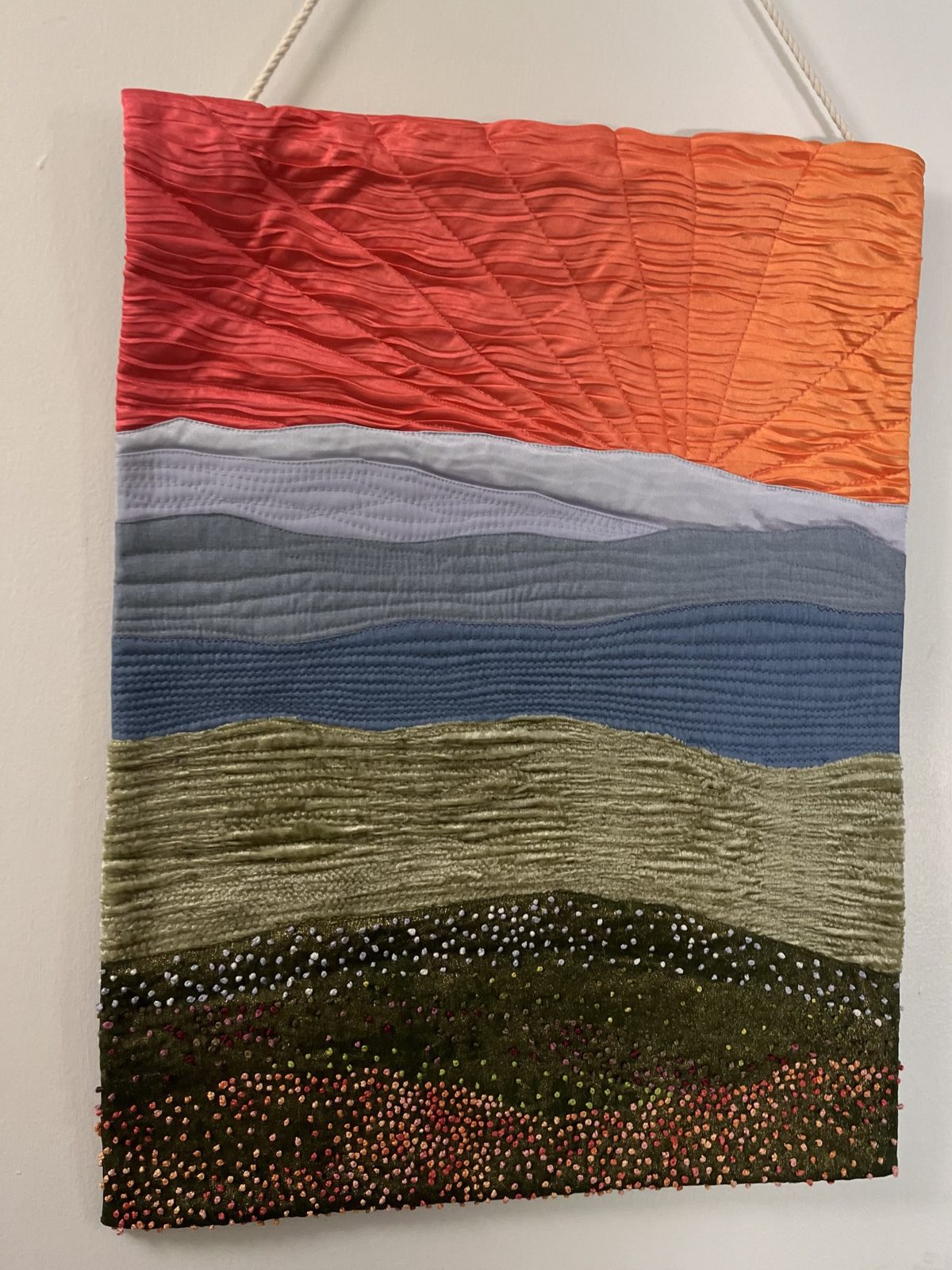 Hanging quilt: orange and coral pleated sunset with radiating quilting, blue ridges, and a dark hill covered in hundreds of French knot wildflowers